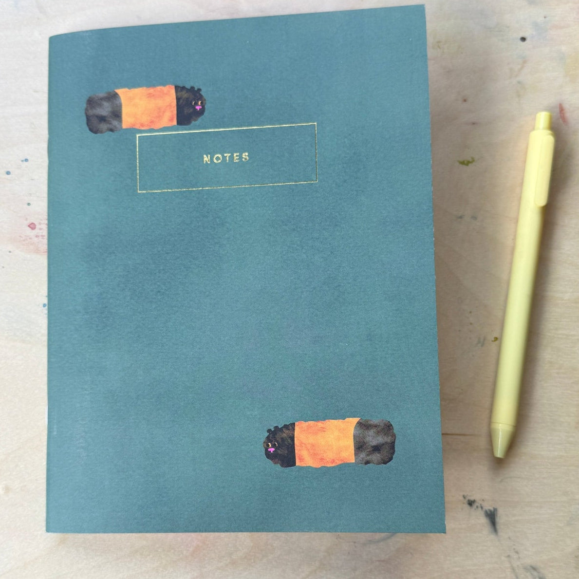 A teal blue notebook with two orange and black cigarette illustrations and a gold-outlined 'NOTES' label, shown on a light wood surface next to a pale yellow pen.